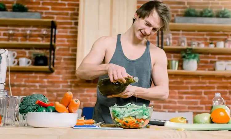 Natural Ways to Support Healthy Hormones A fit man in a grey tank top pouring olive oil into a fresh vegetable salad. He is preparing a healthy meal in a modern kitchen with a brick wall background and fresh produce on the counter for natural ways to support healthy hormones.